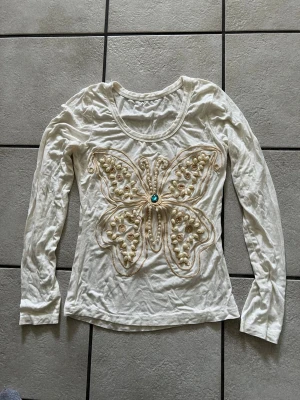 Vit långärmad topp med pärlor storlek S/M - Vit långärmad topp med rund halsringning och stor fjärilsdekor framtill. Fjärilen är dekorerad med pärlor, broderier och en blå sten i mitten. Materialet känns mjukt och lätt, perfekt för dig som gillar unika detaljer.