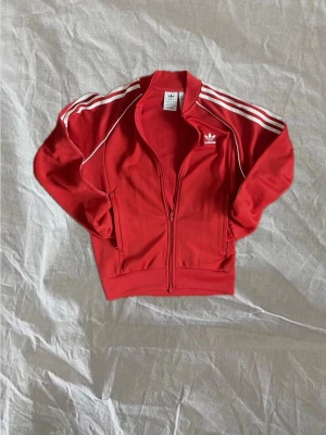 Röd Adidas zip hoodie med vita ränder - Adidas Originals SST track jacket i röd med de klassiska vita ränderna langs armarna. Dragkedja hela vägen upp och två fickor på sidorna. Trevligt skick, inga fläckar, hål eller andra defekter, ser i princip oanvänd ut. Storlek S, sitter som en S ska sitta. Hör av dig om du har frågor!
