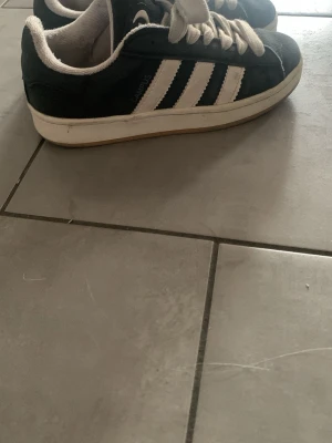 Adidas campus - Svarta sneakers från Adidas Originals med klassiska vita ränder på sidorna och vit sula. Skorna har breda vita skosnören och en logga på plösen. Ovandelen är i mocka och textil, vilket ger en snygg kontrast till de vita detaljerna.