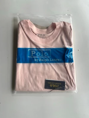 Ljusrosa t-shirt från Polo Ralph Lauren - Snygg ljusrosa t-shirt från Polo Ralph Lauren med klassisk rund hals och liten blå logga broderad på bröstet. Tillverkad i mjuk bomull för skön känsla och normal passform. Perfekt för dig som gillar stilrena och tidlösa plagg.
