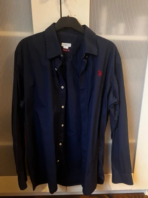 Mörkblå skjorta från U.S Polo Assn. - Klassisk mörkblå skjorta från U.S Polo Assn med röd broderad logga på bröstet. Skjortan har långa ärmar, knappar framtill och en stilren krage. Perfekt för en stilren och avslappnad look.