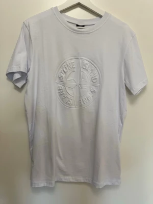 t-shirt med logga - Storlek M/L Vit t-shirt från med logga på bröstet. Klassisk rund hals och korta ärmar. Tillverkad i mjuk bomull för en skön känsla och stilren look.