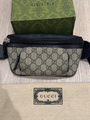 Gucci beige bältesväska äkta  - Snygg bältesväska från Gucci i beige canvas med klassiskt GG-monogram och svarta läderdetaljer. Väskan har justerbart band, dragkedja och en mindre ficka framtill. Perfekt för att bära det viktigaste med stil köpt i Italien 🇮🇹 