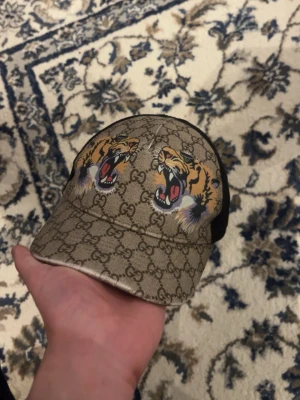 Gucci keps med tigertryck - Snygg keps från Gucci med klassiskt GG-mönster i beige och brunt. Framtill finns två coola tigertryck som verkligen sticker ut. Kepsen har svart mesh på baksidan och justerbart spänne. Perfekt accessoar för att lyfta din stil.