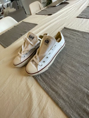 Vita Converse All Star låga sneakers - Klassiska vita Converse All Star sneakers i canvas med lågt skaft och vit gummisula. Skorna har den ikoniska loggan på plösen och detaljer i silver vid snörningen. Perfekta för en avslappnad och stilren look.