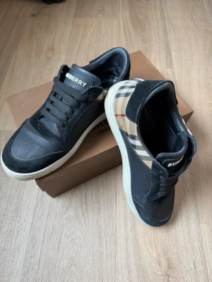 Burberry sneakers med rutigt mönster - Snygga svarta sneakers från Burberry med klassiskt beige rutigt mönster på sidorna. Skorna har vita sulor, svarta skosnören och detaljer i mocka och läder. Perfekta för dig som vill ha en stilren och lyxig look.