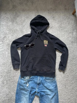 hoodie från Morris - Svart hoodie från Morris med guldfärgad brodyr och brittisk flagga på bröstet. Klassisk känguruficka framtill och justerbar huva med snören. Perfekt för en stilren och avslappnad look.