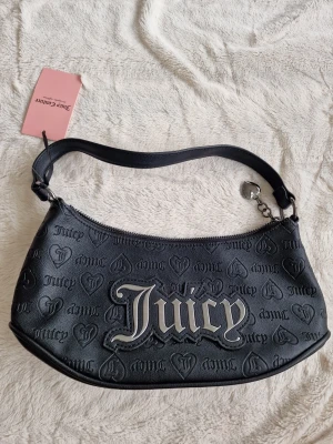 Svart axelväska från Juicy Couture - Cool svart axelväska från Juicy Couture med präglade hjärtan och logotyper över hela väskan. Stor Juicy-logga framtill i silver. Väskan har en kort axelrem och dragkedja upptill. Materialet är konstläder och väskan har en trendig, avlång form.