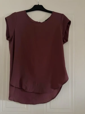 Mörklila blus med rund hals - En stilren mörklila blus med rund halsringning och korta ärmar. Toppen har en lätt rundad nederkant som är något längre bak. Perfekt för en enkel och snygg look.