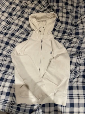 Vit hoodie med dragkedja från Polo Ralph Lauren - Vit hoodie med dragkedja från Polo Ralph Lauren. Klassisk design med huva, långa ärmar och broderad logga på bröstet. Mjuk bomullskänsla och ribbade muddar. Perfekt för en clean och stilren look.