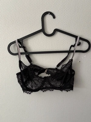 Svart spets-bh med strassband - Säljer en svart bh i genomskinlig spets med dekorativa strassband som axelband. Bh:n har bygel och ett litet hål framtill för extra detalj. Justerbara axelband och hakknäppning bak. Perfekt för dig som gillar en trendig och elegant look.