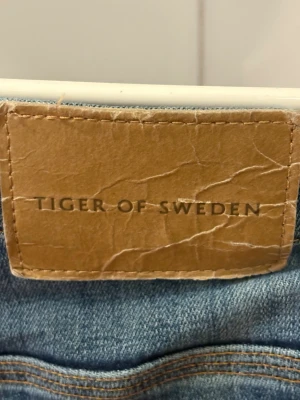 Blå jeans från Tiger of Sweden - Snygga blå jeans från Tiger of Sweden med klassisk femficksdesign och raka ben. Jeansen har en läderpatch med logotyp på bakfickan och är tillverkade i denim. Perfekt för dig som gillar stilrena och tidlösa jeans. Storlek 33/32