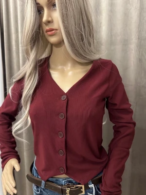 Vinröd ribbad kofta med knappar - Vinröd kofta med ribbad struktur och V-ringning. Långa ärmar och fem matchande knappar framtill. Snygg och enkel att styla till jeans eller kjol. Materialet ser ut att vara bomullsblandning.