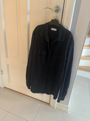 Svart overshirt från Jack and Jones  - Stilren svart overshirt från Jack and Jones med klassisk krage och knäppning framtill. Tillverkad i ett mjukt tyg som känns bekvämt och passar perfekt till lager-på-lager-stil.