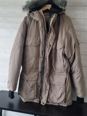 Everest Beige parkasjacka - Snygg beige parkasjacka från Everest med stor huva och grå päls. Jackan har flera stora fickor framtill, dragkedja och knappar samt justerbar midja. Perfekt för kalla dagar och riktigt varm med sitt vadderade material.