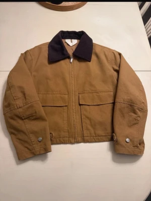 Brun boxy jackafrån Arket - Boxy brun jacka med mörkbrun krage och dragkedja framtill från Arket.                                                   Jackan har två stora fickor fram, knappar vid ärmslut och midja samt en loose passform. Perfekt för dig som gillar en avslappnad stil. 