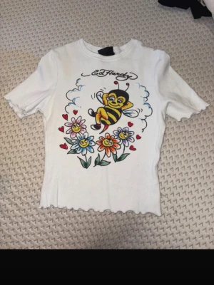 Vit Ed Hardy t-shirt med bi och blommor - Säljer en vit t-shirt från Ed Hardy med ett färgglatt tryck av en tecknad bi, blommor och små röda hjärtan på framsidan. T-shirten har korta ärmar, croppad passform och vågig kant nertill. Perfekt för dig som gillar unika och lekfulla plagg.