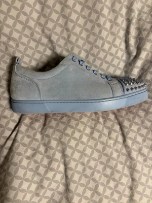 Ljusblå sneakers med nitar - Unika ljusblå sneakers från Christian Louboutin i mocka med coola silverfärgade nitar på tån. Skorna har matchande ljusblå snören och sula, samt diskreta ventilationshål på sidan. Perfekt för dig som vill sticka ut med din stil.