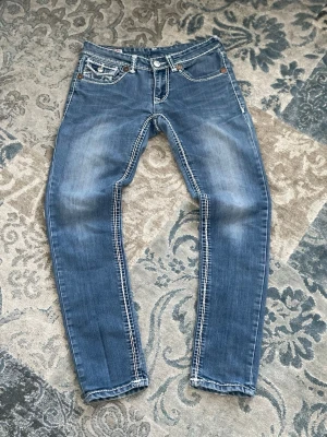 Blåa jeans från True Religion - [FRI FRAKT 🚚]Snygga blå jeans från True Religion med kontrastsömmar och klassisk femficksdesign. Jeansen har en lätt tvättad look och detaljerade sömmar på fickorna. Perfekta för dig som gillar streetstyle och vill sticka ut lite extra.