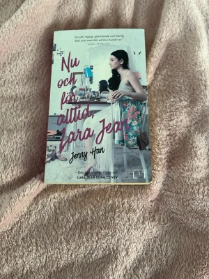 Nu och för alltid, Lara Jean av Jenny Han - Säljer boken 'Nu och för alltid, Lara Jean' av Jenny Han. Omslaget är färgglatt med rosa och turkosa detaljer och visar en tjej vid ett skrivbord. Perfekt för dig som gillar romantisk ungdomslitteratur och contemporary romance.