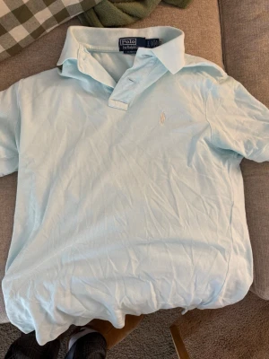 Ljusblå pikétröja från Polo Ralph Lauren - Snygg ljusblå pikétröja från Polo Ralph Lauren med klassisk krage och två knappar vid halsen. Tröjan har det ikoniska broderade logotypen på bröstet och är tillverkad i mjuk bomull. Perfekt för en clean och stilren look. Den blir ljusare ju mer man tvättar den, nypris 2200kr. Det är en S men passar även M