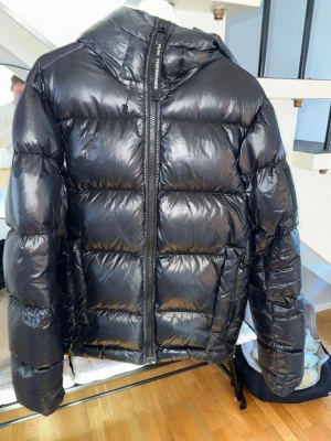 Svart pufferjacka från Peak Performance - Säljer en svart, glansig pufferjacka från Peak Performance med huva och kraftig dragkedja. Jackan har en boxig passform och breda, vadderade paneler som ger en riktigt puffig look. Perfekt för kalla vinterdagar och riktigt snygg till streetwear-stilen.