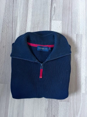 Marinblå halvzip från Henri Lloyd - Snygg marinblå stickad tröja från Henri Lloyd med halvzip och ribbad krage. Tröjan har en diskret röd detalj på insidan av kragen och dragkedjan. Perfekt för dig som gillar klassisk och stilren design. Passar mig som är 163 ≈. 