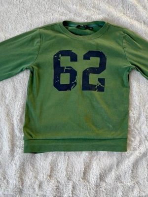 Grön sweatshirt med tryck från H&M - Grön sweatshirt från H&M med stort mörkblått tryck av siffran 62 på bröstet. Tröjan har rund halsringning, långa ärmar och ribbade muddar vid ärmslut och nederkant. Perfekt för en avslappnad och sportig stil.