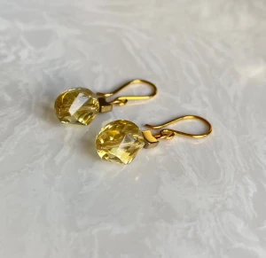 Guldiga örhängen med gul kristall - Snygga örhängen med facetterade, droppformade kristaller i gul nyans och guldfärgade krokar. Kristallerna fångar ljuset och ger ett glittrande intryck. Perfekta för dig som vill ha en färgklick och elegant detalj till din look.