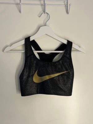 Svart sport-bh från Nike med guldlogga - Snygg svart sport-bh från Nike med en stor guldig Swoosh-logga framtill. Tillverkad i ett glansigt, stretchigt material och har brottarrygg för extra stöd. Perfekt för träning eller gymmet.