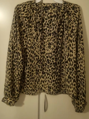 Leopardmönstrad blus med knytband - Snygg blus med leopardmönster i beige och svart. Blusen har långa ärmar och ett dekorativt knytband vid halsen. Tillverkad i ett mjukt, lätt tyg som ger ett avslappnat men trendigt intryck. Perfekt för dig som vill sticka ut med ett djurmönster.