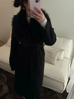 Svart kappa med fuskpäls - Snygg svart kappa med bred krage i fuskpäls och knytskärp i midjan. Lång modell med elegant siluett, perfekt för kalla dagar. Kappan har en lyxig känsla tack vare den fluffiga kragen och passar till många olika stilar.