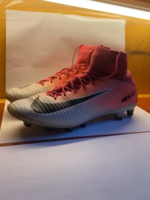 Nike Mercurial rosa/vita fotbollsskor - Snygga Nike Mercurial fotbollsskor i rosa och vitt med svart swoosh på sidan. Skorna har hög strumpkrage för extra stöd och snörning framtill. Tillverkade i syntetmaterial med dobbar för spel på gräs. Perfekta för dig som vill ha stil och prestanda på planen.