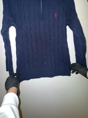 Mörkblå kabelstickad tröja Polo Ralph Lauren - Snygg mörkblå kabelstickad tröja från Polo Ralph Lauren med halv dragkedja och röd broderad logga på bröstet. Tröjan har långa ärmar och ribbade muddar. Perfekt för dig som gillar klassisk och stilren design.