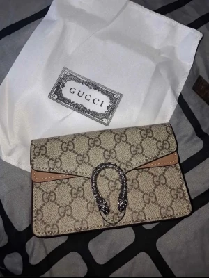 Gucci Dionysus GG Supreme handväska - Säljer en Gucci Dionysus handväska i beige GG Supreme canvas med bruna detaljer och silverfärgad kedja. Väskan har det klassiska GG-mönstret och ett snyggt spänne med ormhuvuden framtill. Perfekt för dig som gillar lyxiga accessoarer och trendig design.