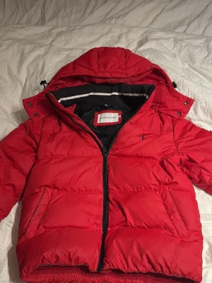 Röd pufferjacka från Calvin Klein  - Snygg röd pufferjacka från Calvin Klein med huva och dragkedja framtill. Jackan har två stora fickor, svart innerfoder och elastisk nederkant. Perfekt för kalla dagar och har en sportig vibe med diskret logga på bröstet.
