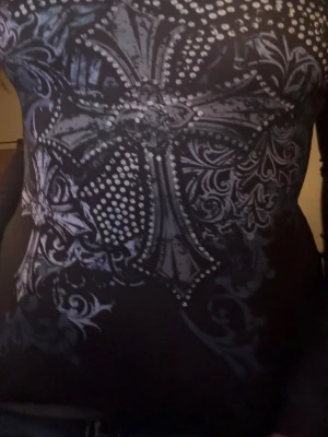 Svart långärmad topp med tryck - Säljer en svart långärmad topp med stort, detaljerat tryck i grått och vitt framtill. Toppen har en slim passform och är tillverkad i ett stretchigt material som sitter snyggt på kroppen. Perfekt för dig som gillar edgy och unika plagg. Vit på insidan, sportigt material. 