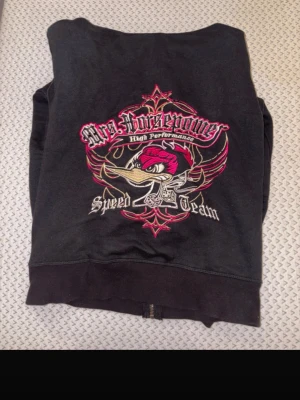 Svart Mrs Horsepower hoodie - Cool svart hoodie från Mrs Horsepower med stort broderat motiv av en arg fågel och texten 'High Performance Speed Team' i rosa, vitt och gult på ryggen. Mindre logga på bröstet och dragkedja framtill. Perfekt för dig som gillar streetwear och unika detaljer.