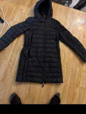 Svart lång dunjacka från Parajumpers - Snygg svart dunjacka från Parajumpers med huva och dragkedja framtill. Jackan är lång, har två sidofickor med dragkedja och ett tydligt Parajumpers-märke på ärmen. Perfekt för kalla dagar och riktigt skön att bära.