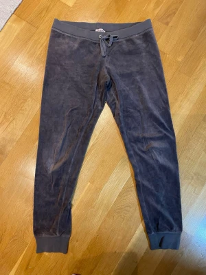 Bruna velour mjukisbyxor från Juicy Couture  - Bruna mjukisbyxor i mjuk velour med resår och dragsko i midjan från Juicy Couture. Byxorna har muddar vid bensluten och två bakfickor med knappdetaljer. Perfekta för chill dagar eller när du vill vara extra bekväm.