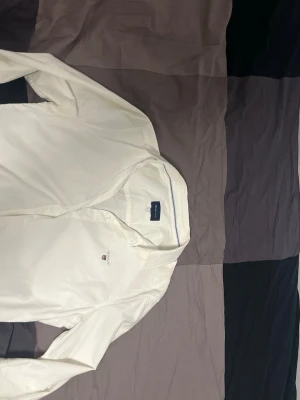 Vit skjorta från Gant med knappar - Klassisk vit skjorta från Gant med diskret logga broderad på bröstet. Skjortan har lång ärm, knappar framtill och är tillverkad i bomull. Perfekt för en clean och stilren look.