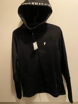 Svart hoodie från Peak Performance M  - Splitterny Snygg svart hoodie från Peak Performance med dragkedja framtill och två fickor med dragkedja. Logga på bröstet och texten 'Peak Performance' tryckt på huvan. Tillverkad i ett mjukt och stretchigt material som känns sportigt och modernt.