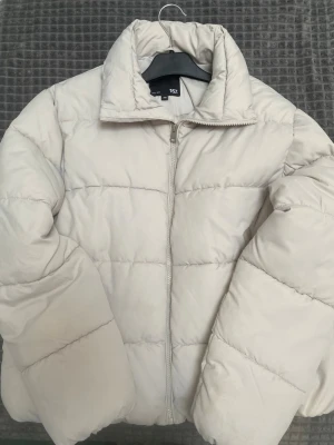 Beige pufferjacka med hög krage - Stilren beige pufferjacka från H&M med hög krage och dragkedja framtill. Jackan har en quiltad design och breda kanaler som ger en fluffig look. Perfekt för kalla vinterdagar och enkel att matcha med olika outfits.