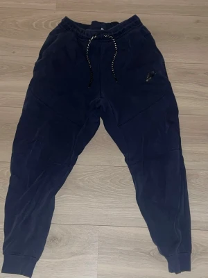 Mörkblå Nike mjukisbyxor - Säljer ett par mörkblå mjukisbyxor från Nike med dragsko i midjan och ribbade benslut. Byxorna har en ficka med dragkedja på baksidan och ett diskret Nike-logga på framsidan. Perfekta för chill eller träning.
