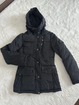 Svart pufferjacka Rockandblue - Säljer en svart pufferjacka från Rockandblue med huva och flera fickor framtill. Jackan har dragkedja, tryckknappar och Rockandblue-logga på ärmen. Perfekt för kalla dagar och riktigt skön att bära. Klassisk quiltad design och snygga detaljer.