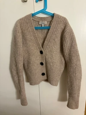 Beige ribbstickad kofta från &otherstories - Mysig beige ribbstickad kofta från &otherstories med v-ringning och tre stora svarta knappar framtill. Koftan har lång ärm. 