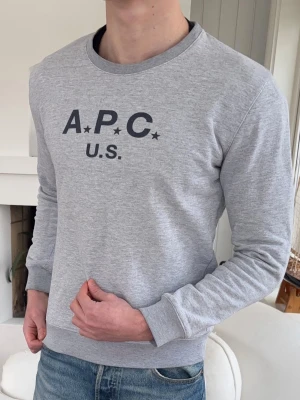 Grå sweatshirt från A.P.C. - Snygg ljusgrå sweatshirt från A.P.C. med svart tryck på bröstet där det står 'A.P.C. U.S.' och två stjärnor. Tröjan har rund halsringning, långa ärmar och ribbade muddar. Perfekt till jeans för en avslappnad look.