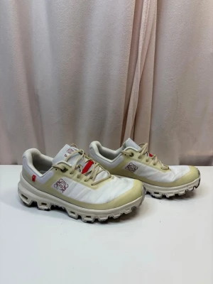 Loewe x On Running  - Unika vita sneakers från Loewe x On Running med beige detaljer och röd logga på sidan. Skorna har chunky sula med CloudTec-teknologi och är tillverkade i textil och syntet. Perfekta för dig som vill sticka ut med stilrena och sportiga detaljer.