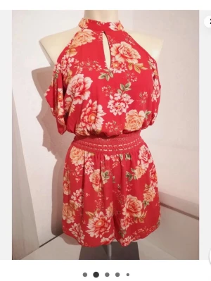 Röd blommig halterneck jumpsuit - Röd blommig playsuit från New Yorker. Cold shoulder, knytning i nacken och markerad midja. Helt ny med prislapp kvar. Storlek 34.