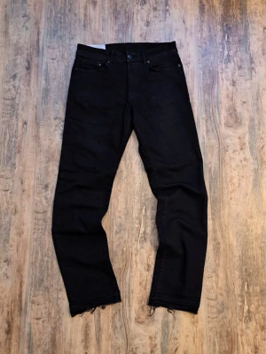 Svarta raka jeans med rå kant - Säljer ett par svarta jeans med rak passform och råa, fransiga benslut. Klassisk femficksmodell med knappgylf och bälteshällor. Jeansen har en stilren look och passar perfekt till en avslappnad outfit.